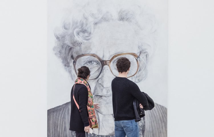 A woman and a man looking at a large monochrome sketch of an elderly woman in an art gallery